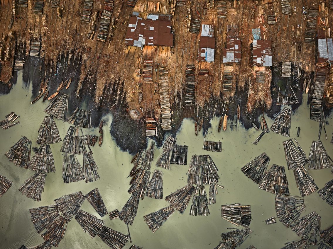 Edward Burtynsky, Sawmills #1, Lagos, Nigeria, 2016 from the Anthropocene series. Photograph. ©Edward Burtynsky. Courtesy of Weinstein Hammons Gallery, Minneapolis / Nicholas Metivier Gallery, Toronto.  Nigeria is a global hotspot for pristine forest loss. In previously unspoiled rainforests, loggers cut down trees, dumping them in the river to float downstream to the sawmills of Lagos. There, logs are resized into lumber for firewood and furniture.   The process despoils the environment in both locations. Rainforest destruction erodes soils, pollutes streams, and turns fertile land to desert. Milling generates sawdust and muddies Lagos’ waterways.   Burtynsky makes the devastation visible, spurring us to question how we manage earth’s resources.