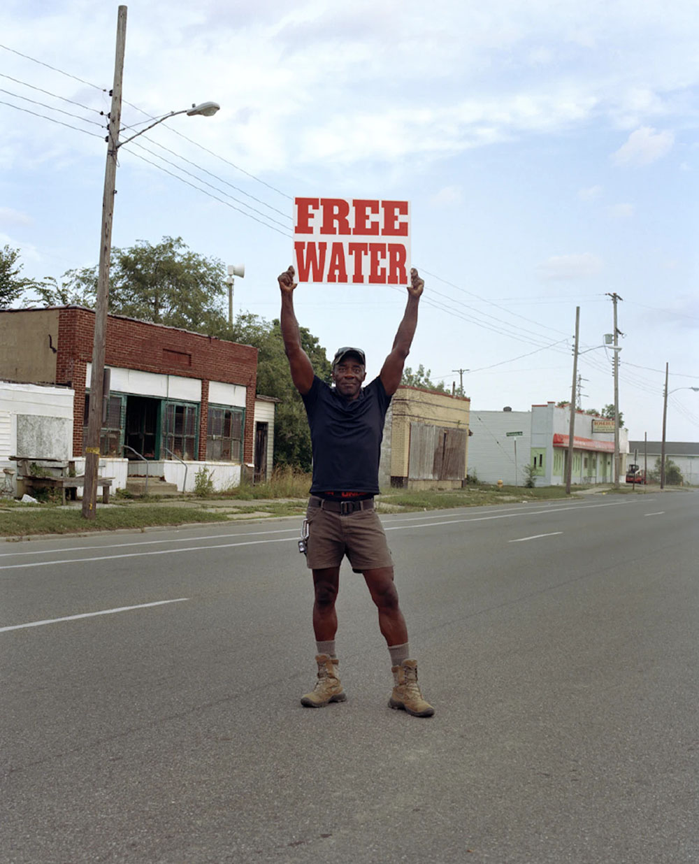 Moses West Holding a “Free Water” Sign on North Saginaw Street Between East Marengo Avenue and East Pulaski Avenue, Flint, Michigan, 2019. ©2022 LaToya Ruby Frazier. Courtesy of the artist and Gladstone Gallery.